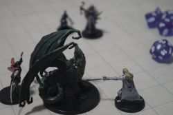 Photo of miniature figures on a grid map. Largest central figure is a dragon. 4 smaller figures surrounding it. Blue polyhedral dice off to the side.